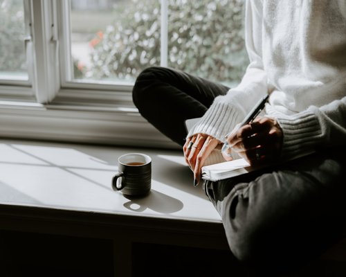 Person relaxing with a cup of chai reading a book near window